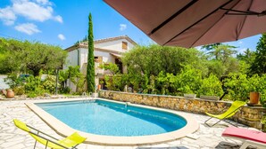 Una piscina con borde de piedra, rodeada de vegetación y un edificio al fondo.