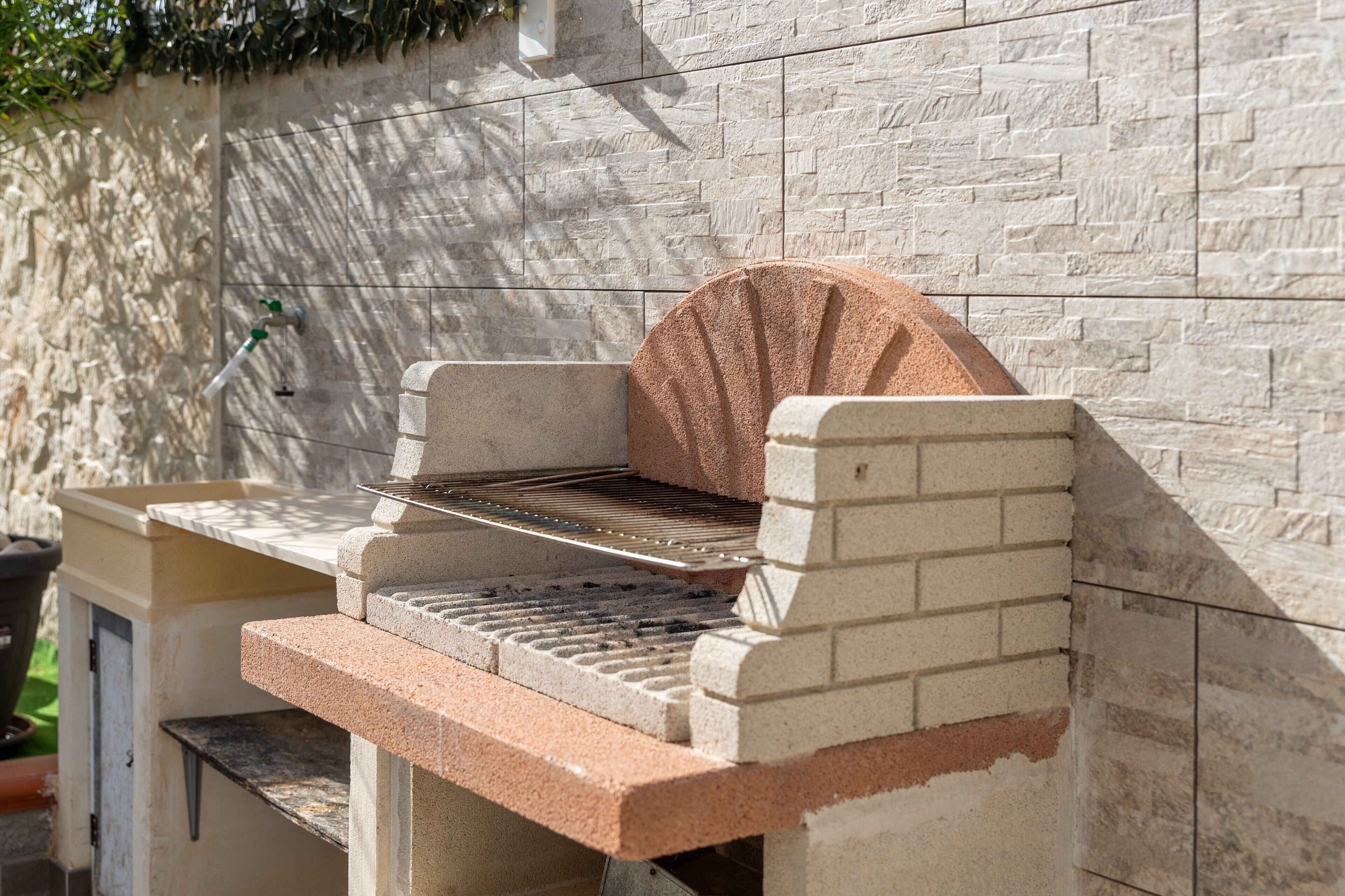 A stone barbecue grill with a metal grate, set against a textured stone wall.