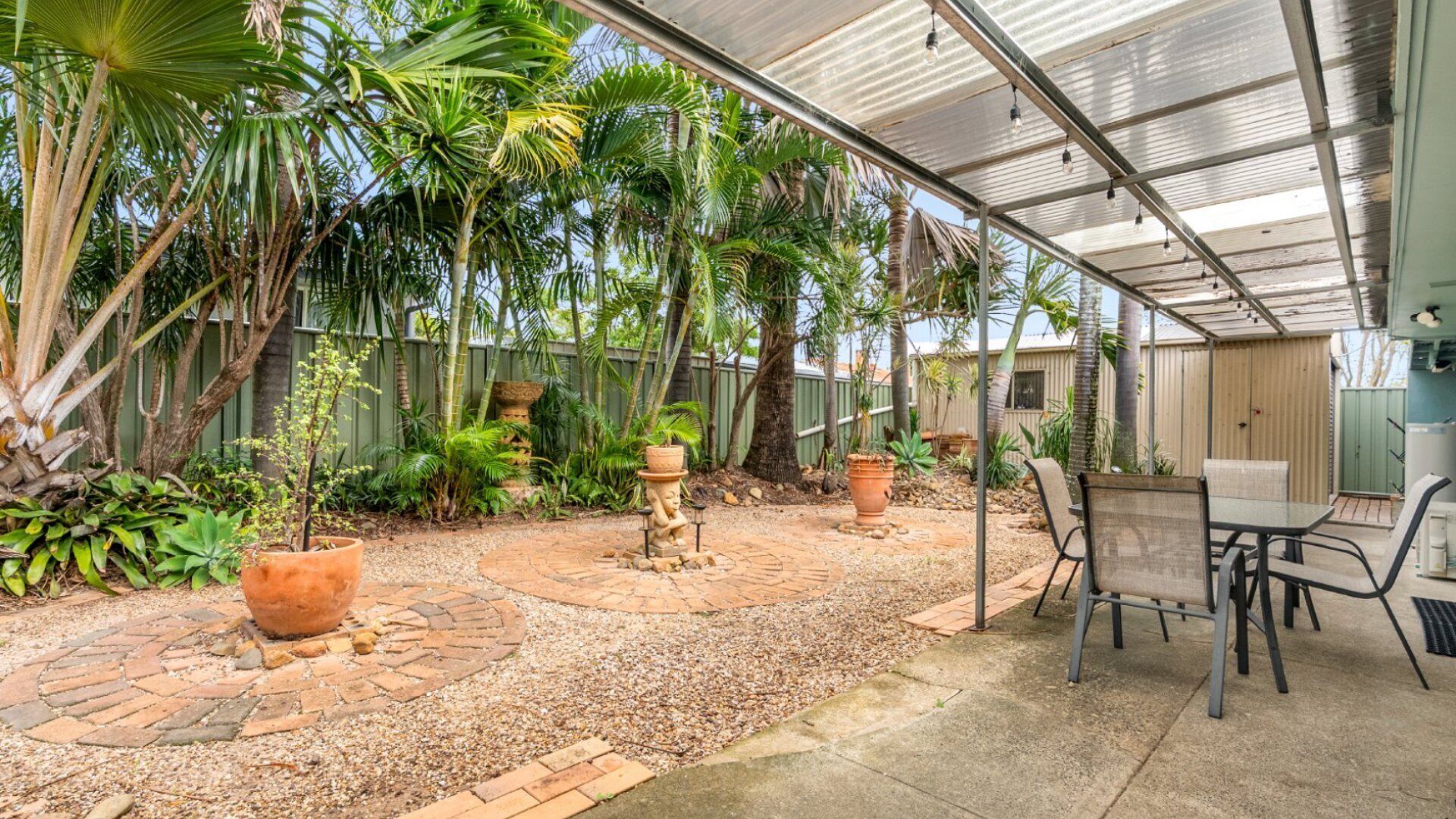 A covered patio area with a table and chairs, surrounded by potted plants and a gravel path.