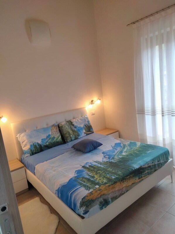 A bedroom with a bed, nightstands, a wall-mounted light, and a window with curtains.