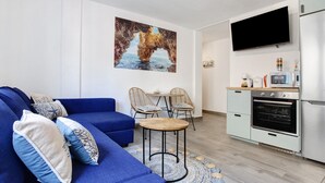 Un salon moderne avec un canapé bleu, deux fauteuils, une petite table, un coin cuisine avec un téléviseur et un grand tableau accroché au mur.