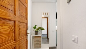 Un couloir avec une porte en bois, une plante en pot, une console blanche avec des tiroirs et un mur blanc avec un interrupteur.