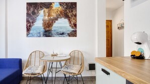 Une pièce comprenant un coin repas avec une table et deux chaises en osier, un canapé bleu, un comptoir de cuisine avec un appareil à gaufres et un téléviseur fixé au mur.