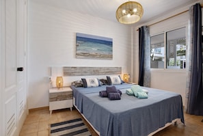 Une chambre à coucher avec un lit, une table de chevet, une lampe, un tableau et une fenêtre avec des rideaux.