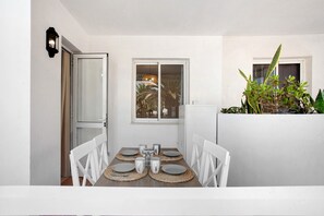 Un coin repas avec une table dressée pour quatre personnes, des chaises blanches, une fenêtre donnant sur un beau paysage et une plante sur la droite.
