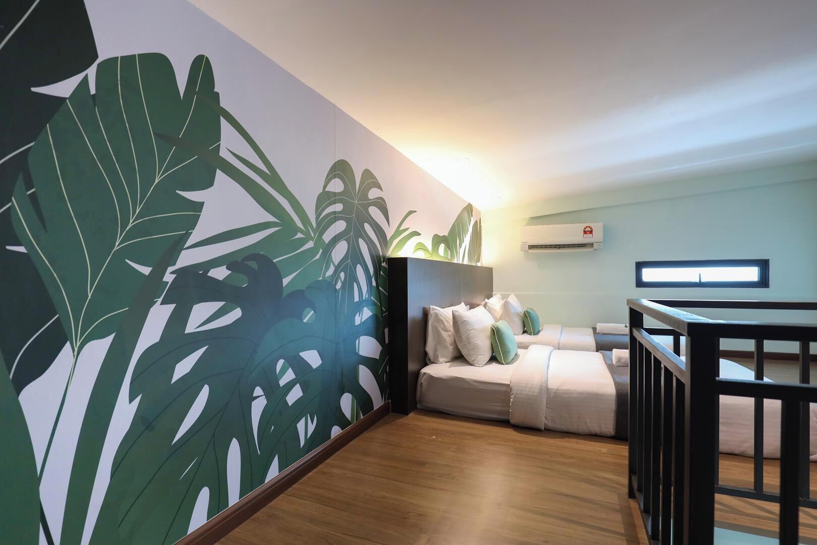 A hotel room with a large tropical leaf mural, a bed with pillows, a wooden floor, and a window.