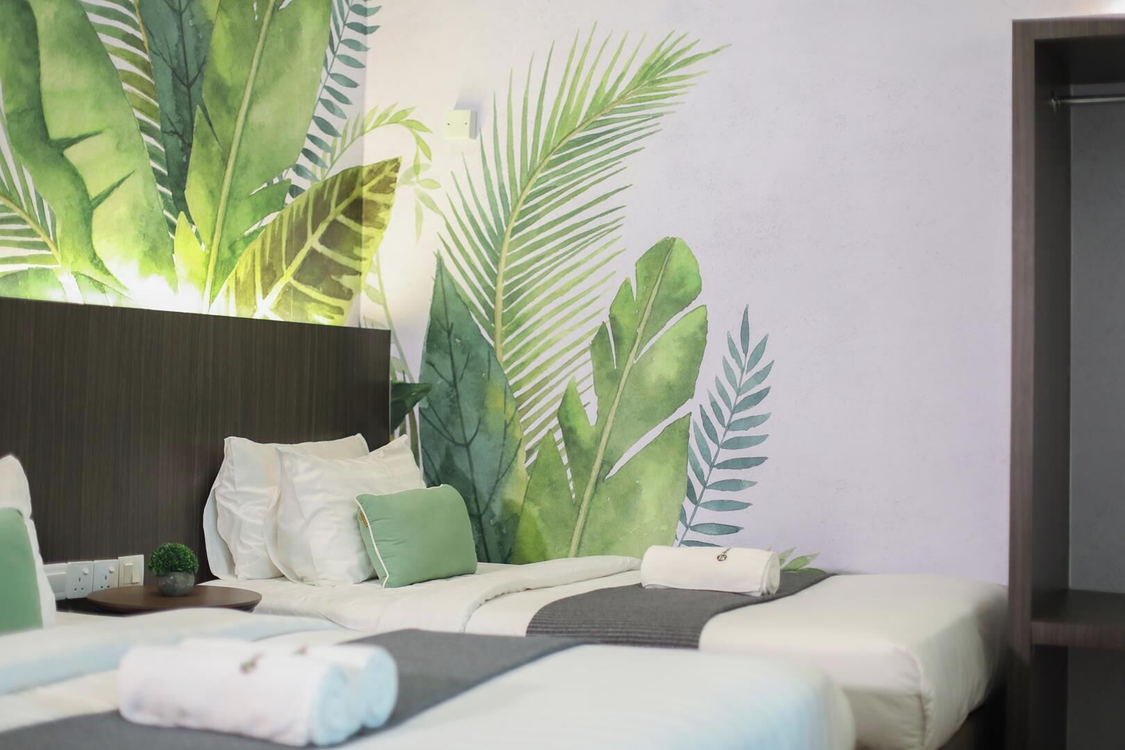 Quarto de hotel com cama, almofadas, uma pequena mesa com uma planta e uma parede com mural de folhagem tropical.
