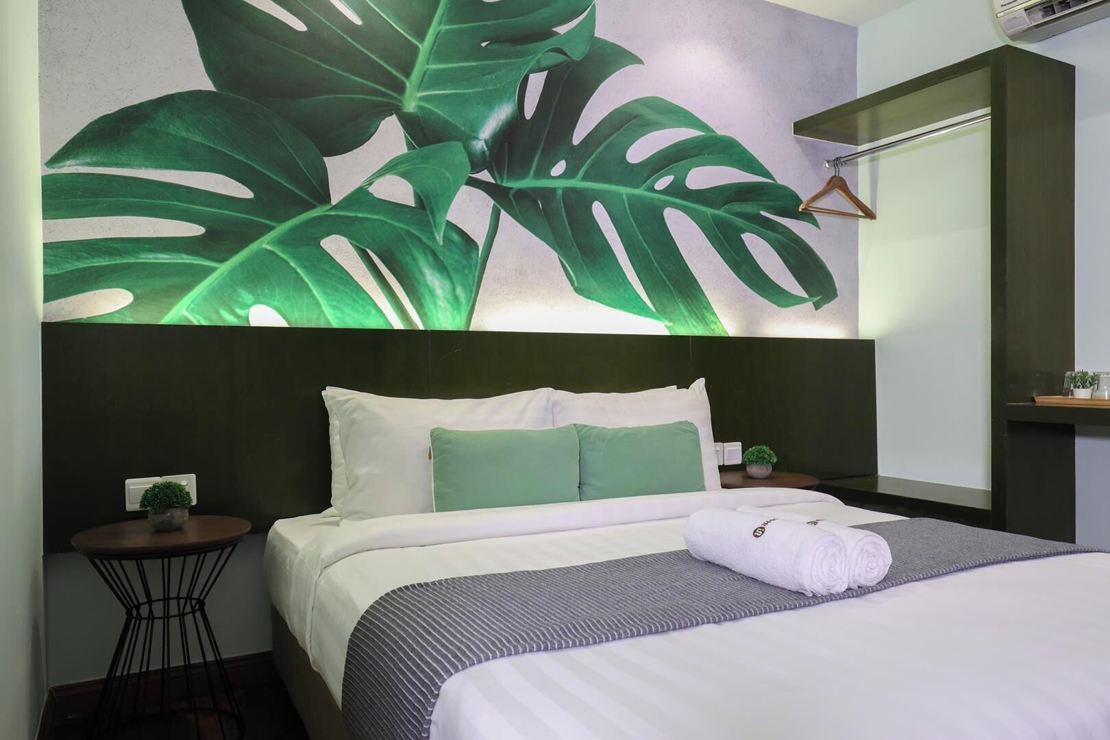 Habitación de hotel con cama, mesita de noche, unidad de aire acondicionado de pared y un mural grande con una hoja verde.