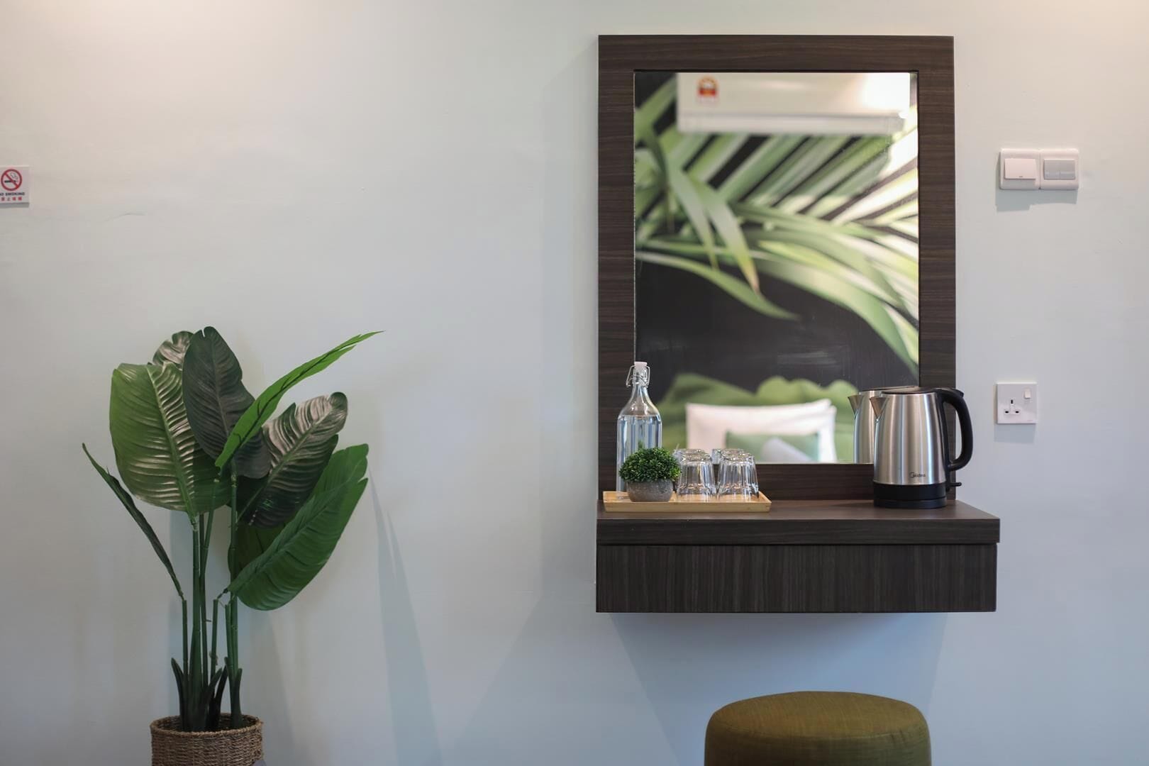 A wall-mounted mirror with a plant and a tray of glasses and a kettle on a dark wooden shelf.