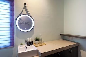 Un baño con un lavabo blanco, un espejo con luz, una planta pequeña en maceta y artículos de aseo en la encimera.
