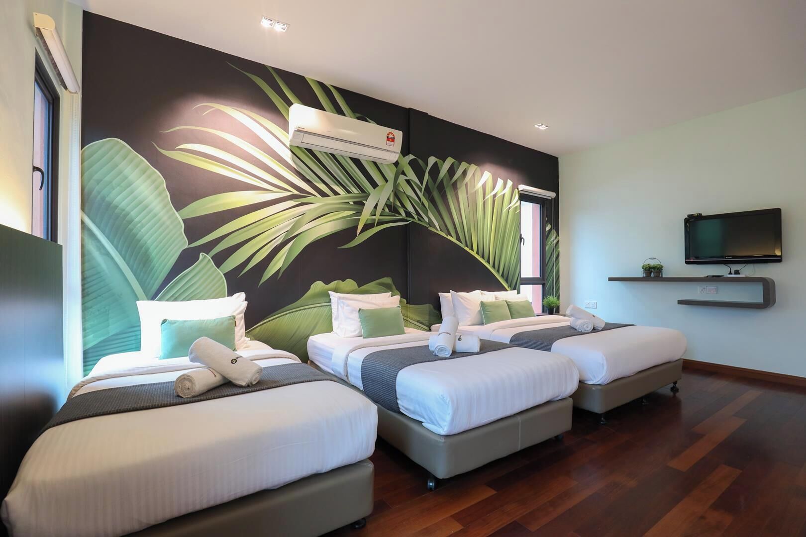 A hotel room with three beds, a large wall mural, an air conditioning unit, a television, and a small shelf with a plant.