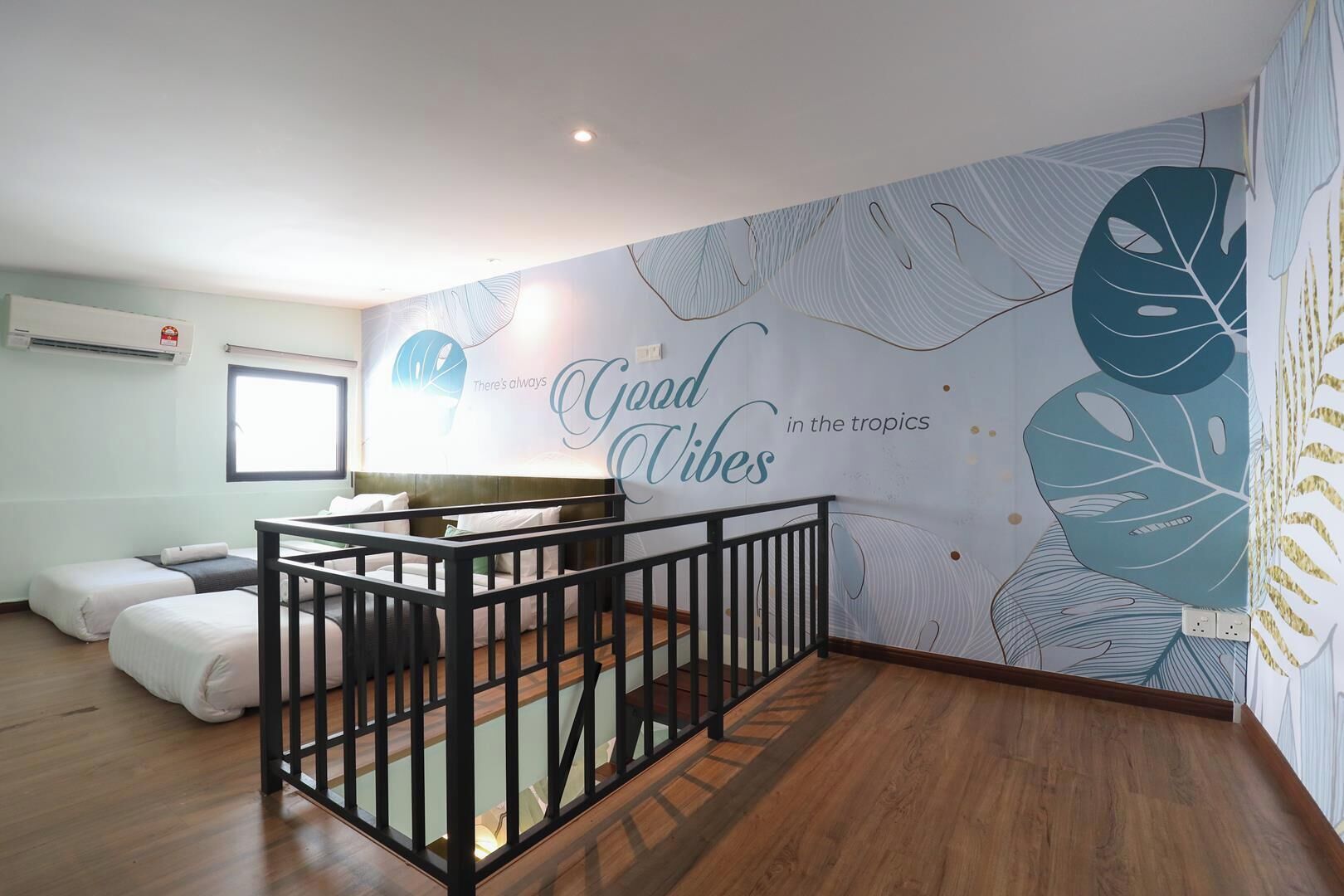 A room with a wooden floor, a black railing, a bed, a desk, and a wall with tropical leaf patterns and the text 'Good Vibes in the tropics'.