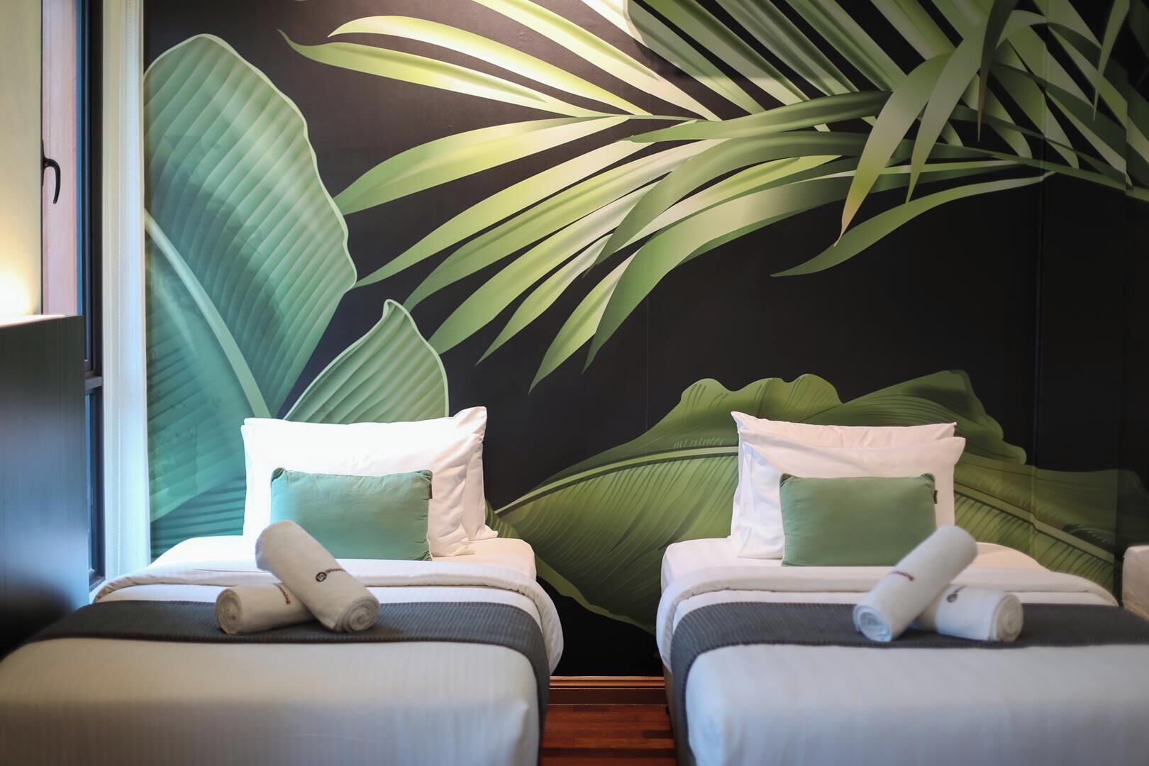 Habitación de hotel con dos camas, cada una con almohadas y toallas enrolladas, frente a una pared con un mural de hojas.