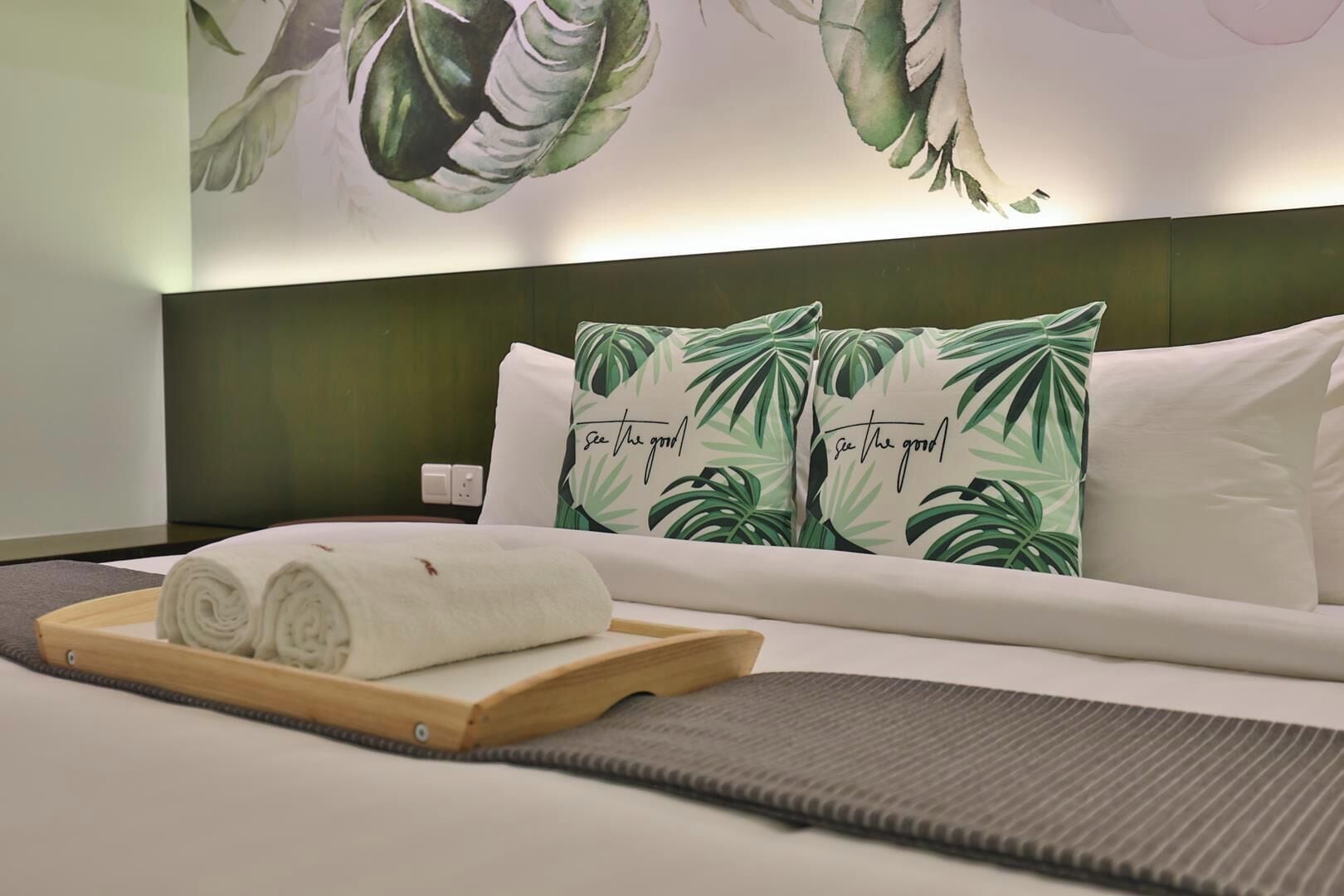 Une chambre d’hôtel avec un lit, des oreillers ornés d’un motif de feuilles tropicales, un plateau avec des serviettes roulées et un mur décoré d’une grande œuvre d’art représentant des feuilles.