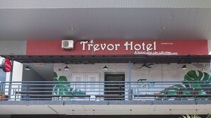 Exterior del Trevor Hotel con fachada roja y blanca, un área de balcón cubierta y un cartel publicitario del hotel.