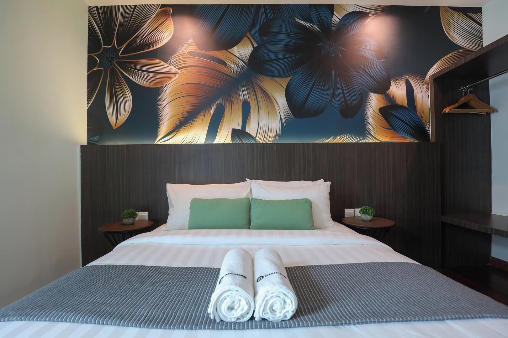 Ett hotellrum med en stor säng, två kuddar, en prydligt vikt handduk, ett litet bord med en växt och en väggmålning.