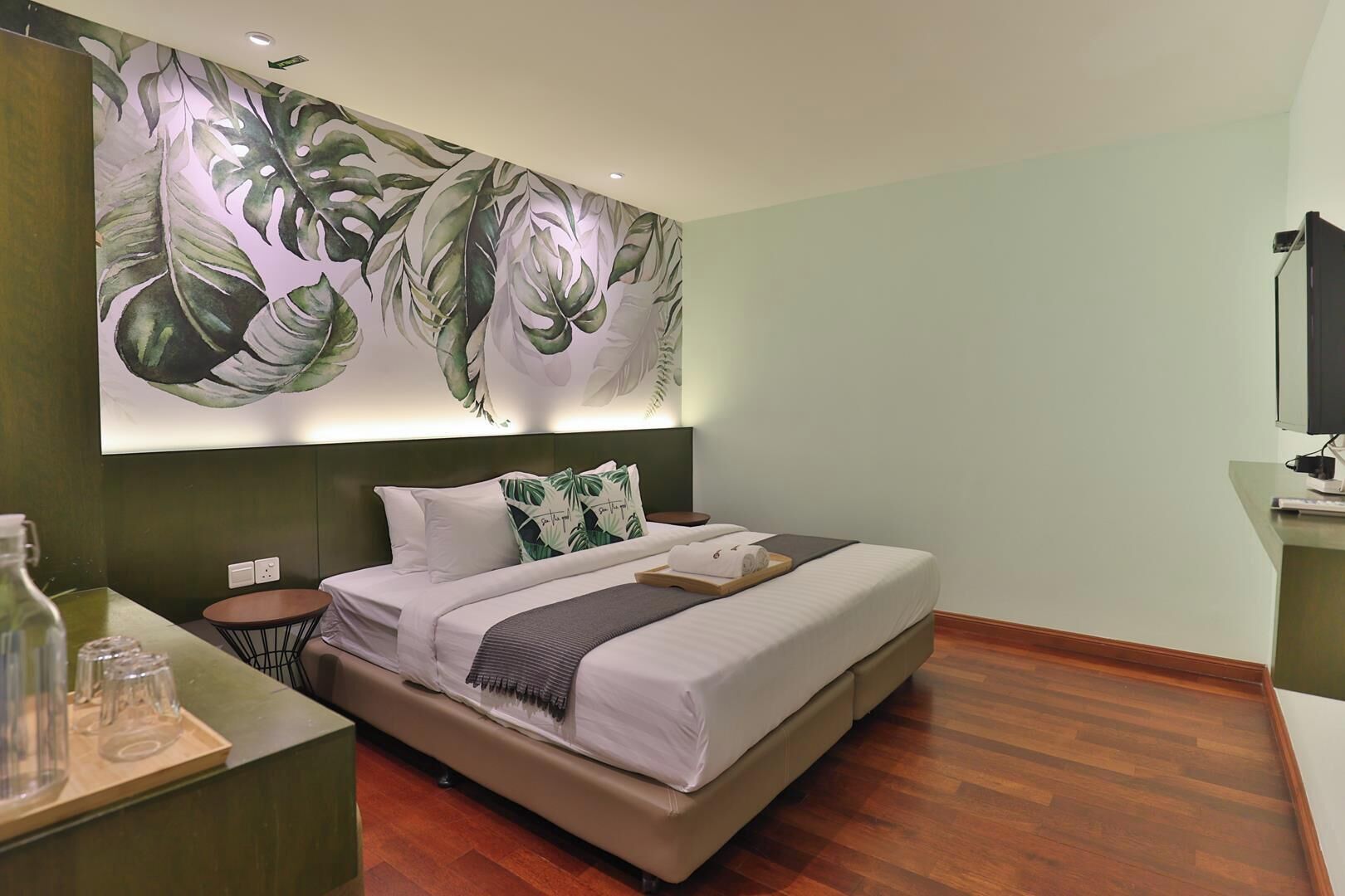 Quarto moderno com uma cama grande, uma mesa de cabeceira, televisão fixada na parede e um mural com folhagem tropical.