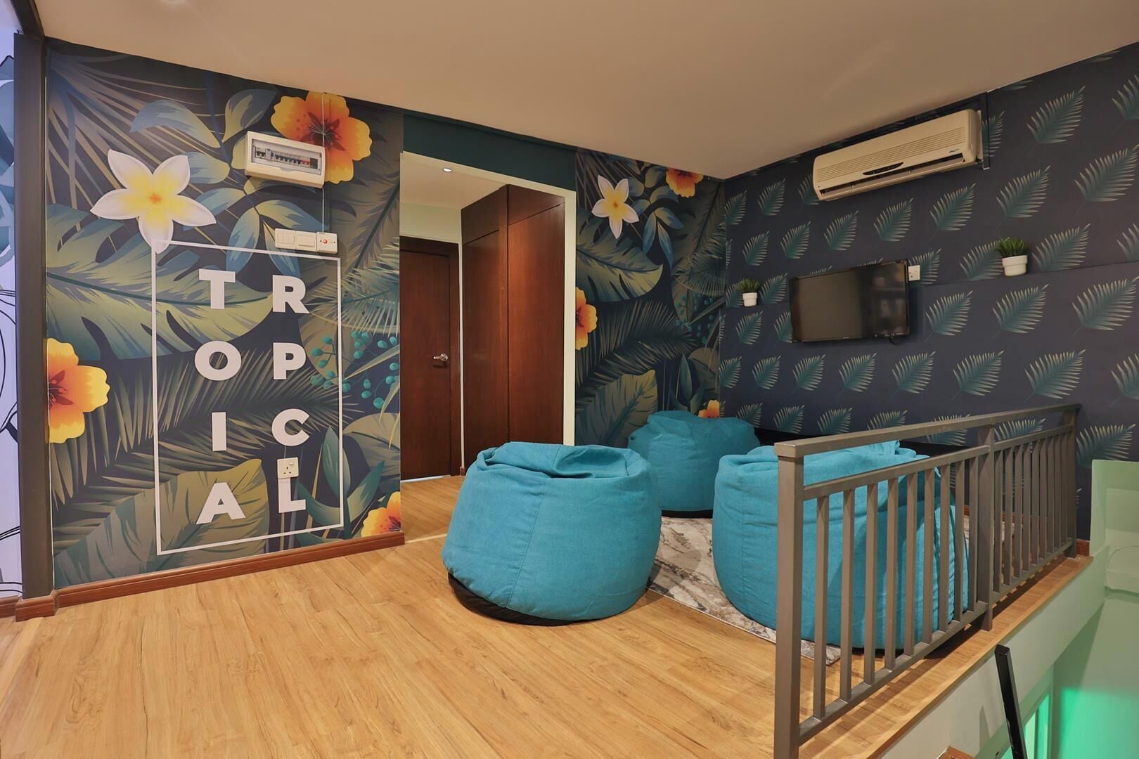 A room with a wooden floor, a blue bean bag chair, a crib with a grey railing, and a wall with a tropical floral wallpaper.
