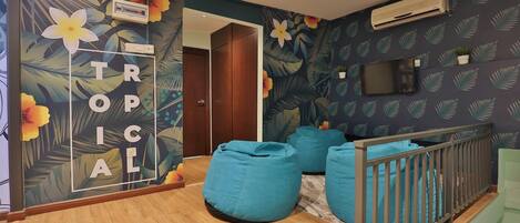 A room with a wooden floor, a blue bean bag chair, a crib with a grey railing, and a wall with a tropical floral wallpaper.
