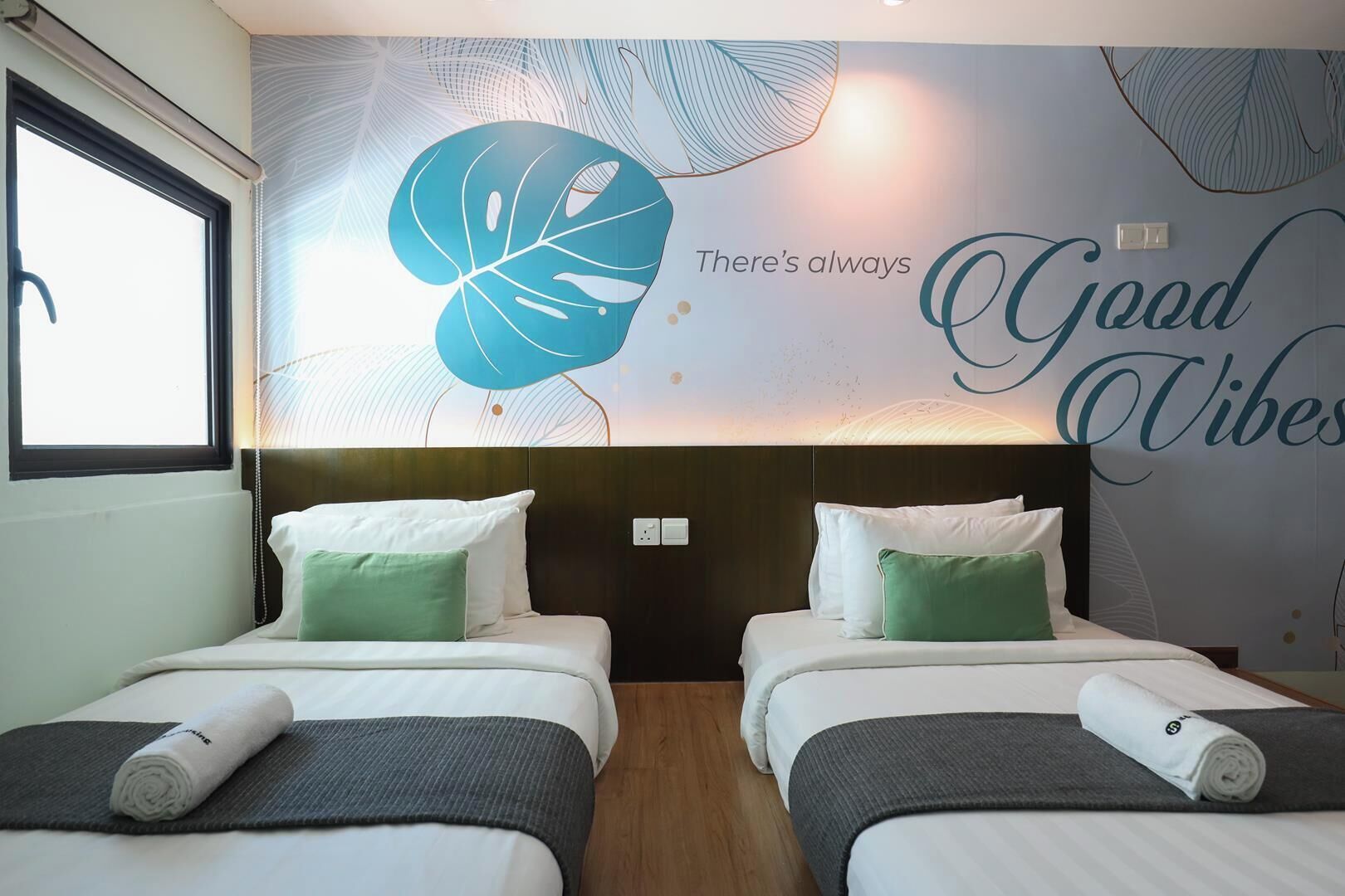 A hotel room with two beds, a large wall mural with a leaf design, and the text 'There's always Good Vibes'.