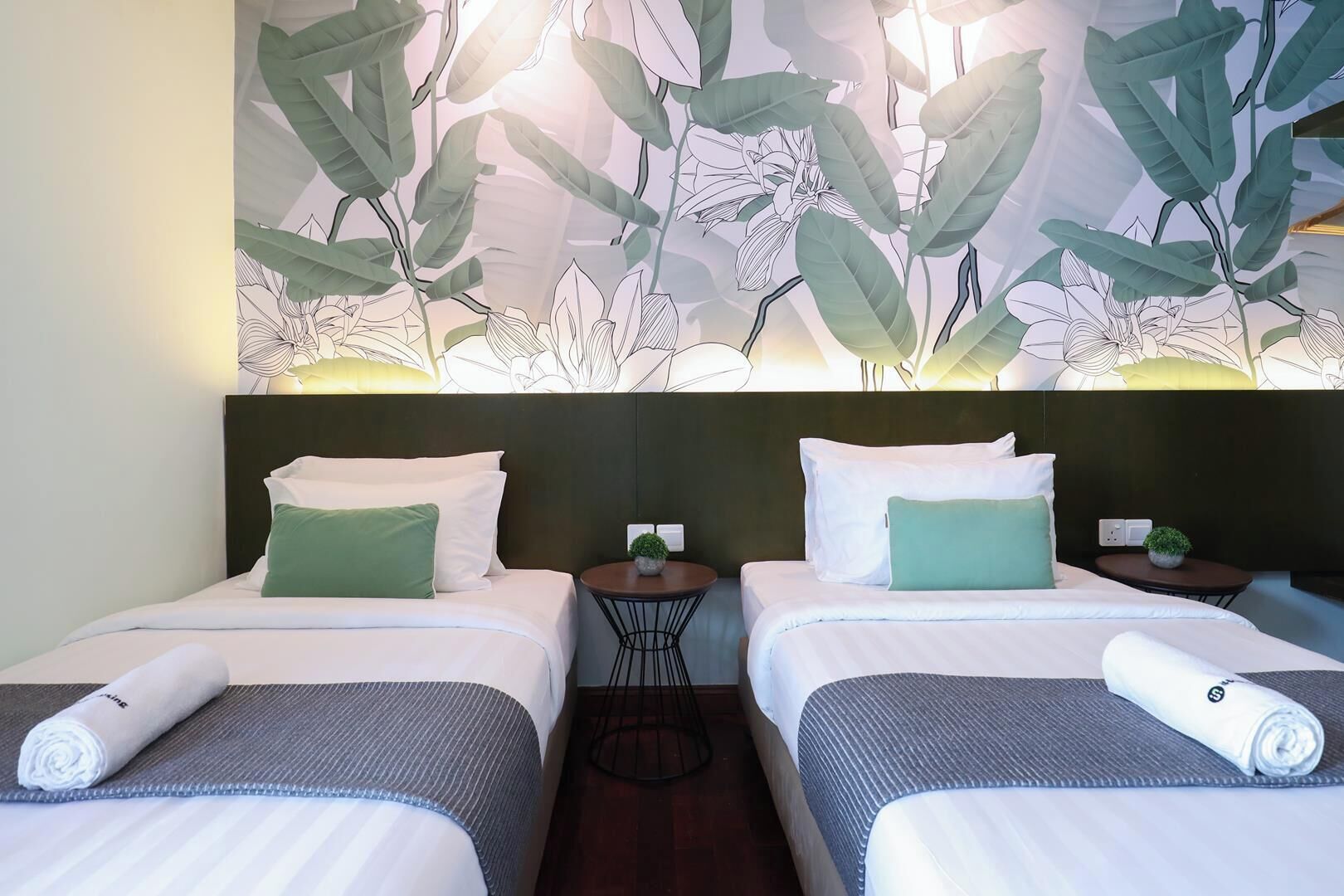 Een hotelkamer met twee bedden, elk voorzien van wit beddengoed en groene kussens, een donker hoofdeinde en een wanddecoratie met bladeren.