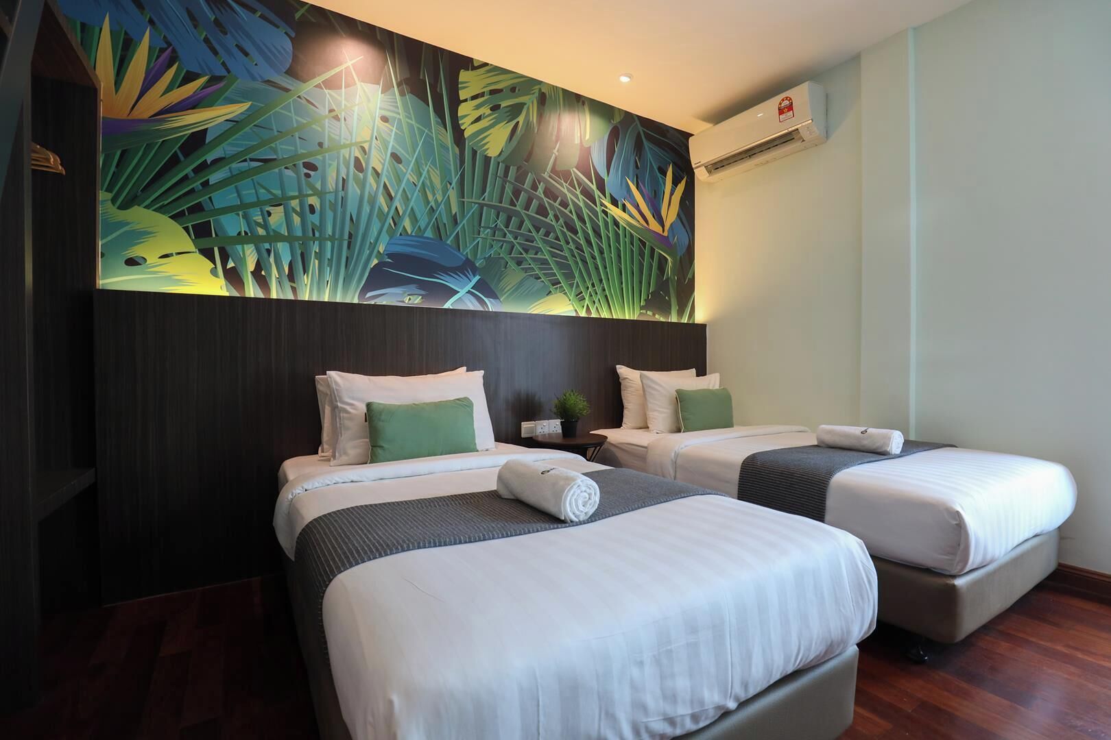 Habitación de hotel con dos camas, un mural tropical grande, una mesita con una planta y un equipo de aire acondicionado.