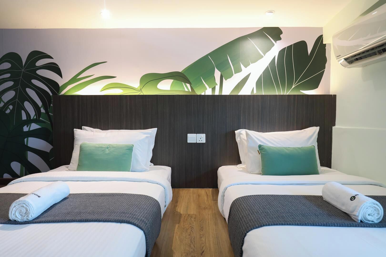 Une chambre d’hôtel avec deux lits, une grande fresque murale représentant des feuilles tropicales et un appareil de climatisation.
