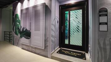 A hotel entrance with a modern design, including a large green plant, a door with a striped pattern, and a sign indicating a walk-in guest service area.