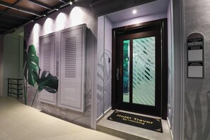 A hotel entrance with a modern design, including a large green plant, a door with a striped pattern, and a sign indicating a walk-in guest service area. - Trevor Hotel by I Housing (Malacca City)