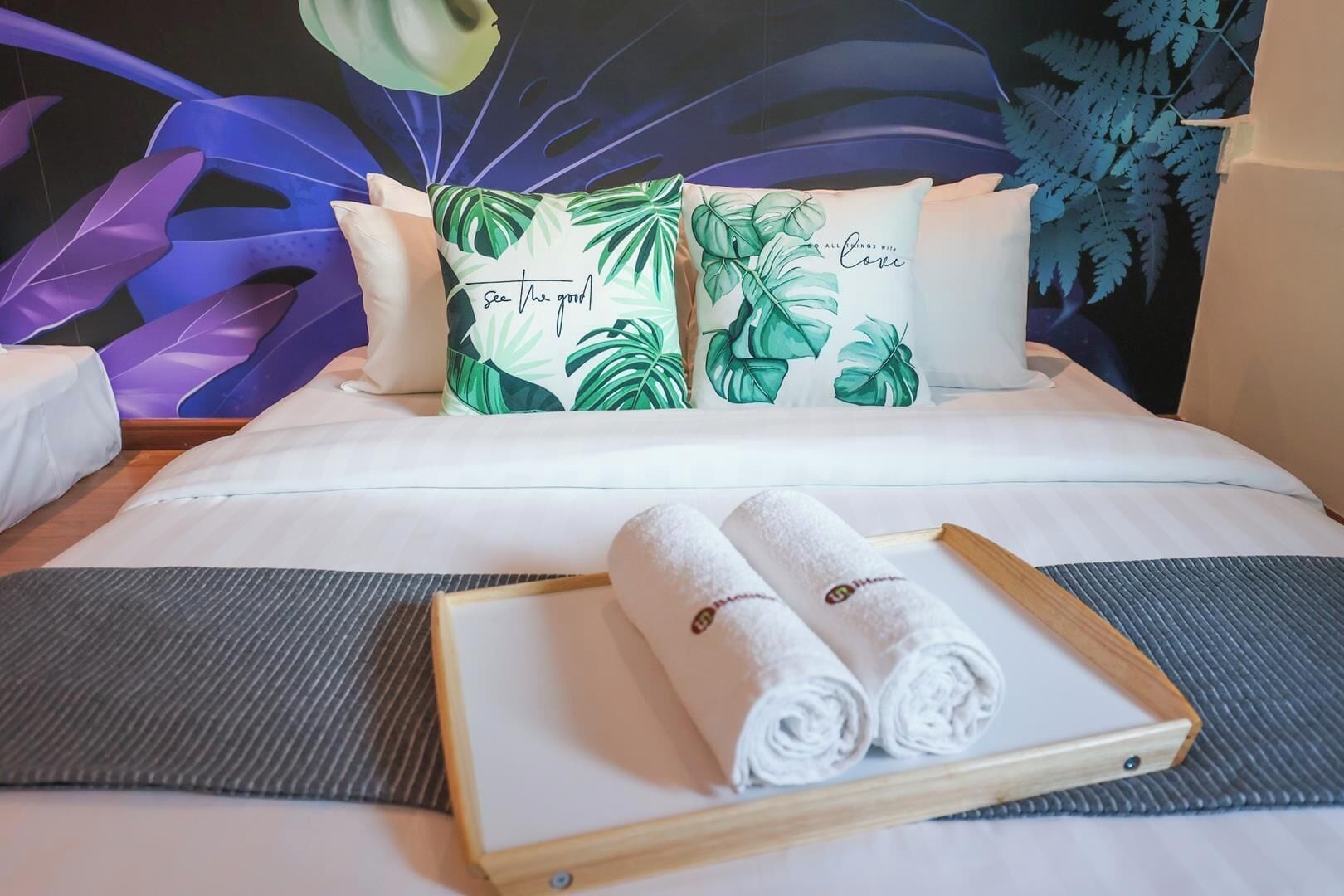 A neatly made bed with white linens, two pillows with a tropical leaf pattern, and a pair of rolled towels on a tray.
