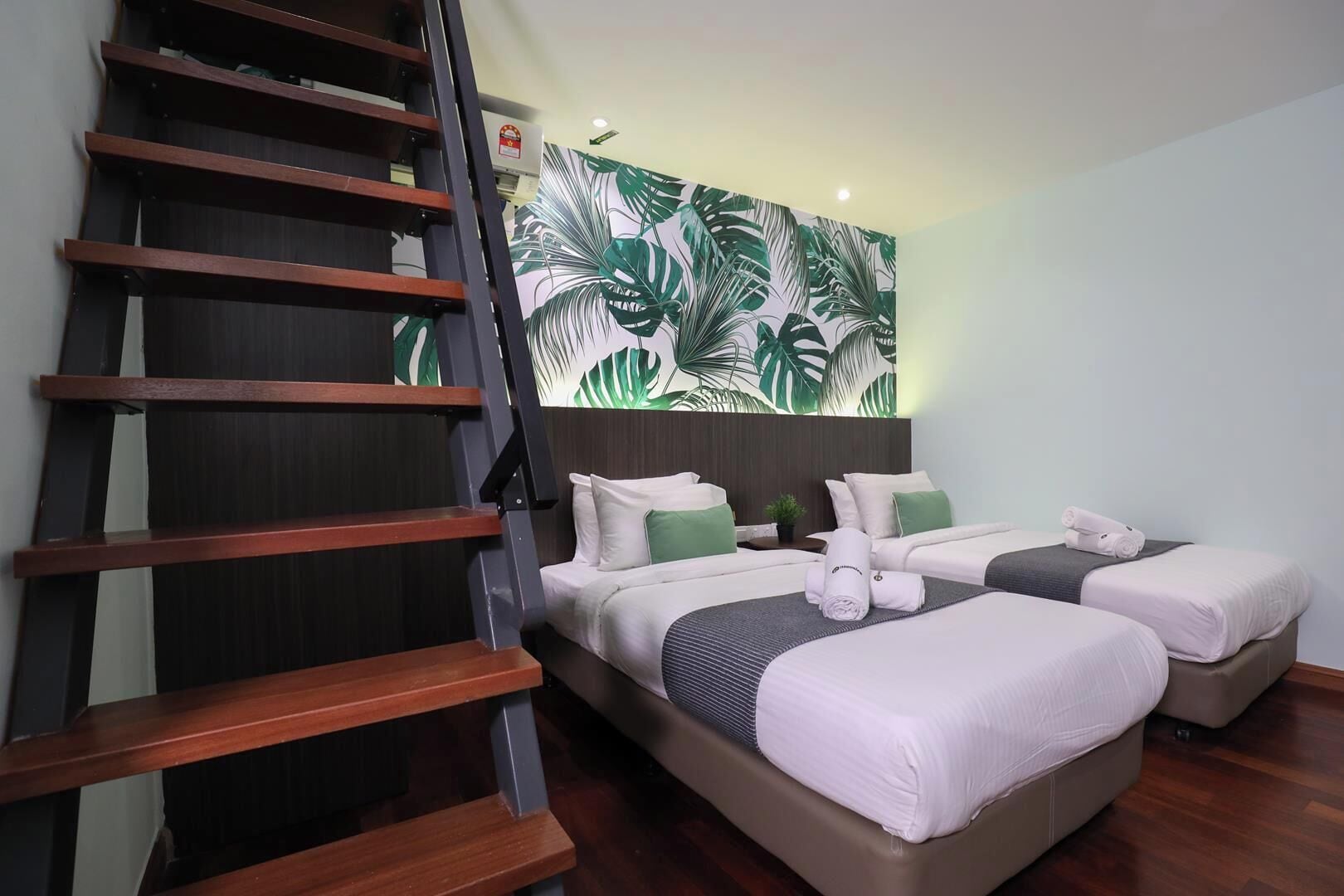 A hotel room with a staircase, two beds with white bedding, green pillows, and a tropical leaf wall mural.