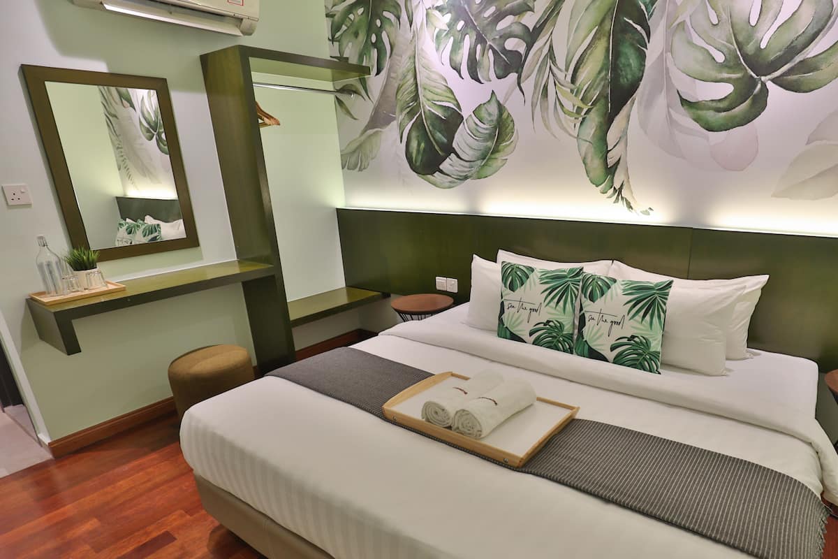 A neatly made bed with white linens and green pillows, a bedside table with a tray of glasses and a bottle, and a large mirror above the bed.