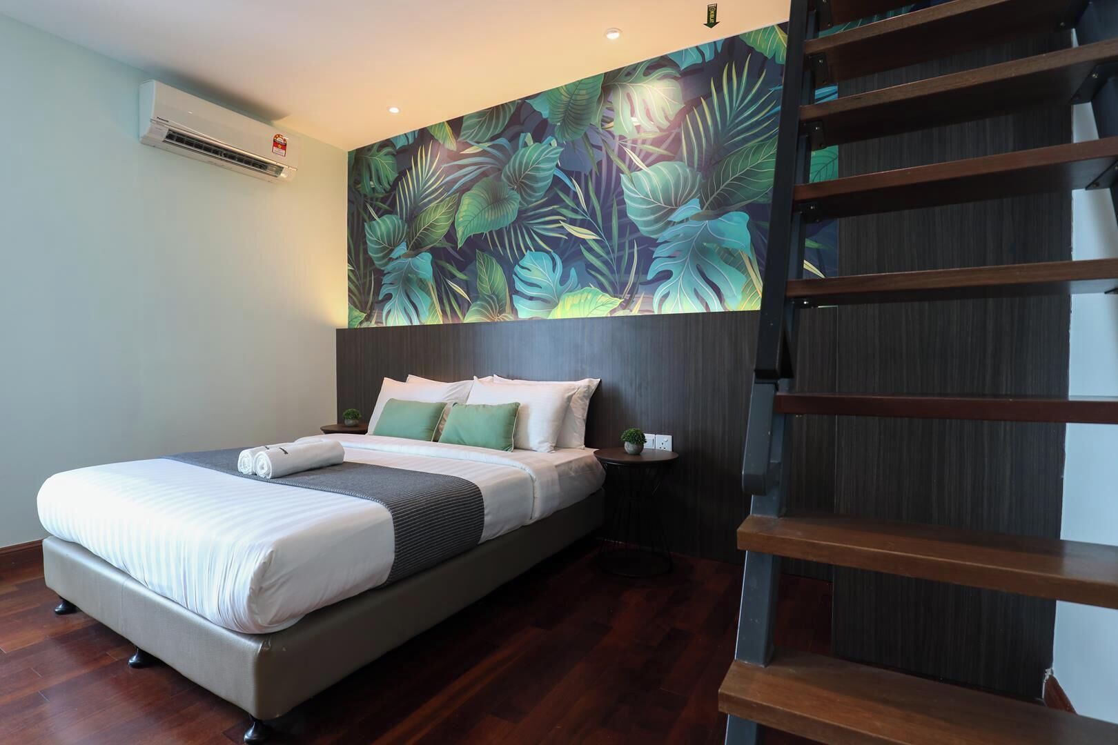 A hotel room with a bed, a nightstand, a wall-mounted air conditioner, and a large tropical leaf mural.