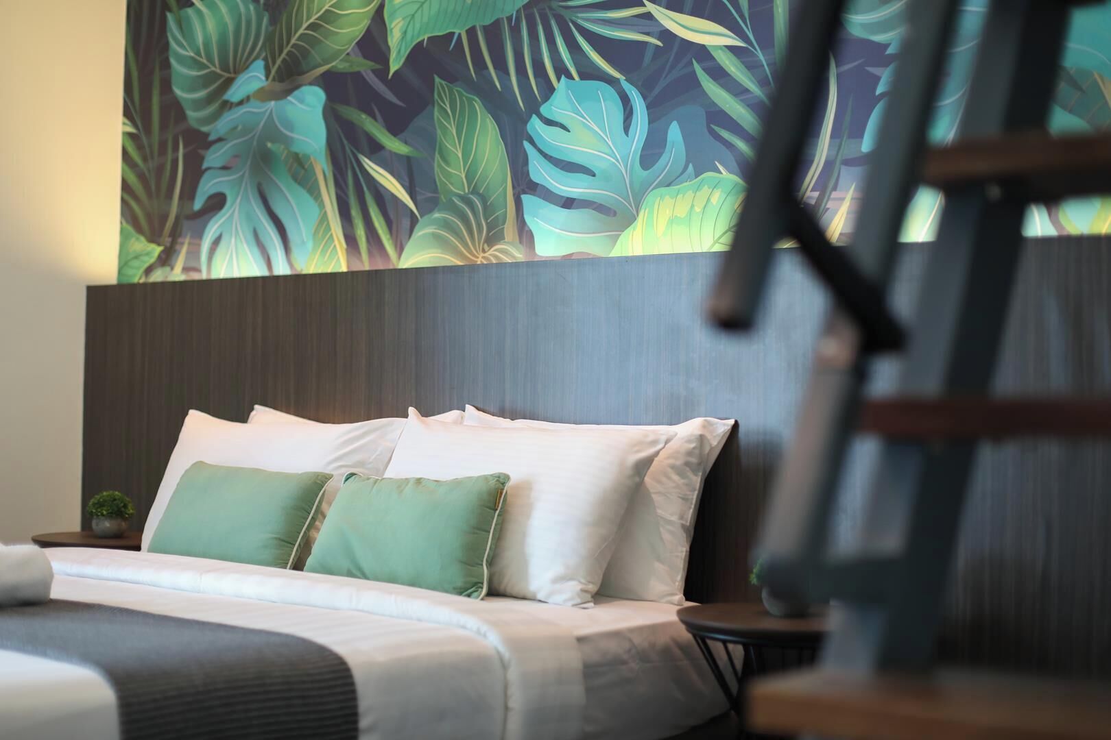 Ein Hotelzimmer mit Bett, Kissen, einem Nachttisch, einer kleinen Pflanze und einer Wand mit tropischem Blattmuster.