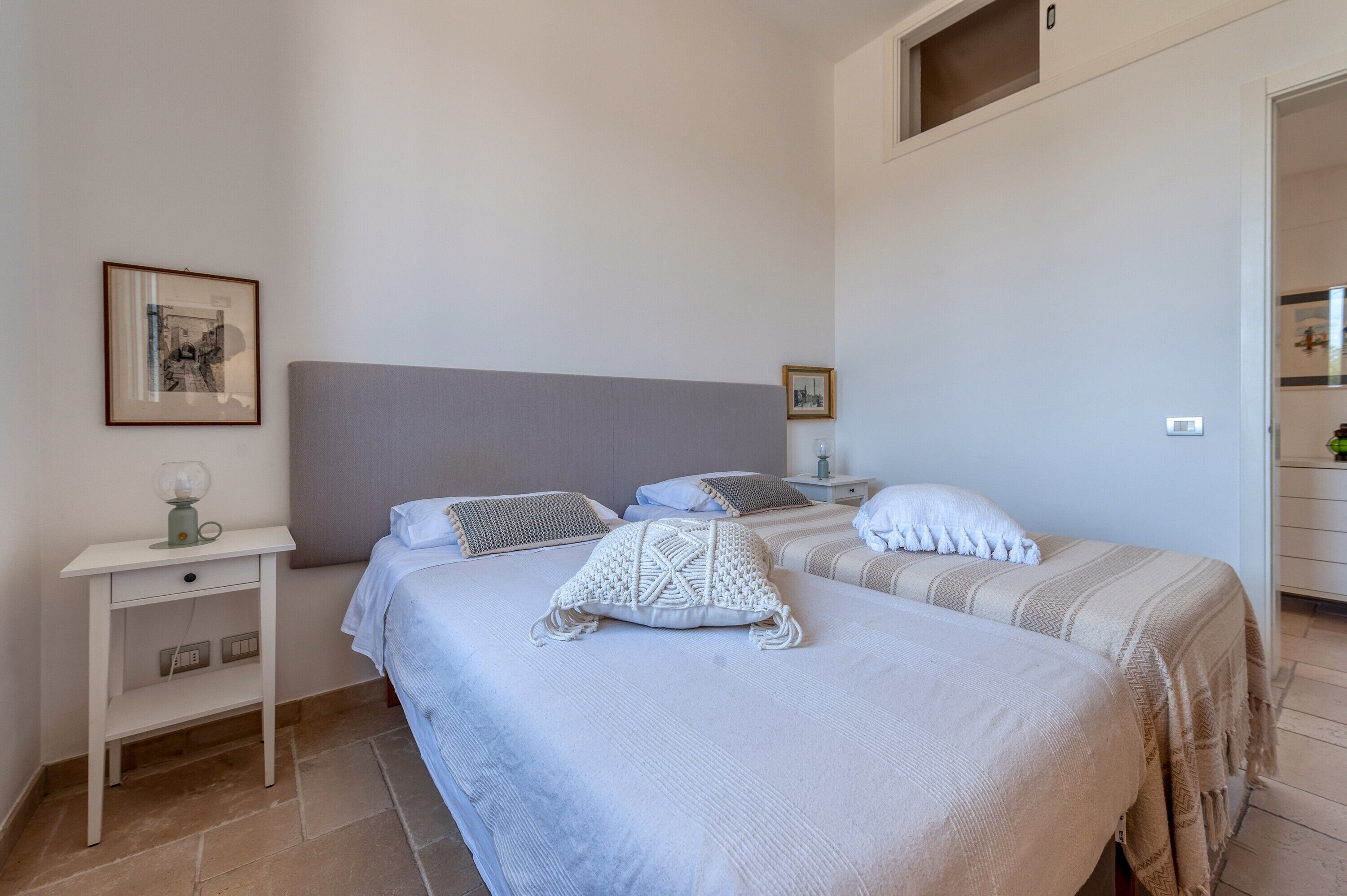 A bedroom with two beds, a nightstand, a framed picture, and a small window.