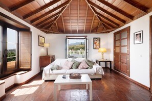 Una habitación con techo de madera, un sofá blanco con cojines, una mesita y una ventana con persianas, con vistas al exterior.