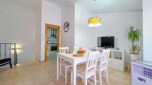 Un comedor con una mesa y sillas blancas, un televisor, una planta en maceta y una cocina visible al fondo.