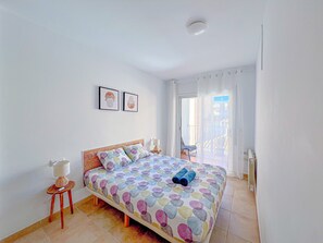 A bedroom with a bed, a chair, a small table, a radiator, and a sliding glass door leading to a balcony.