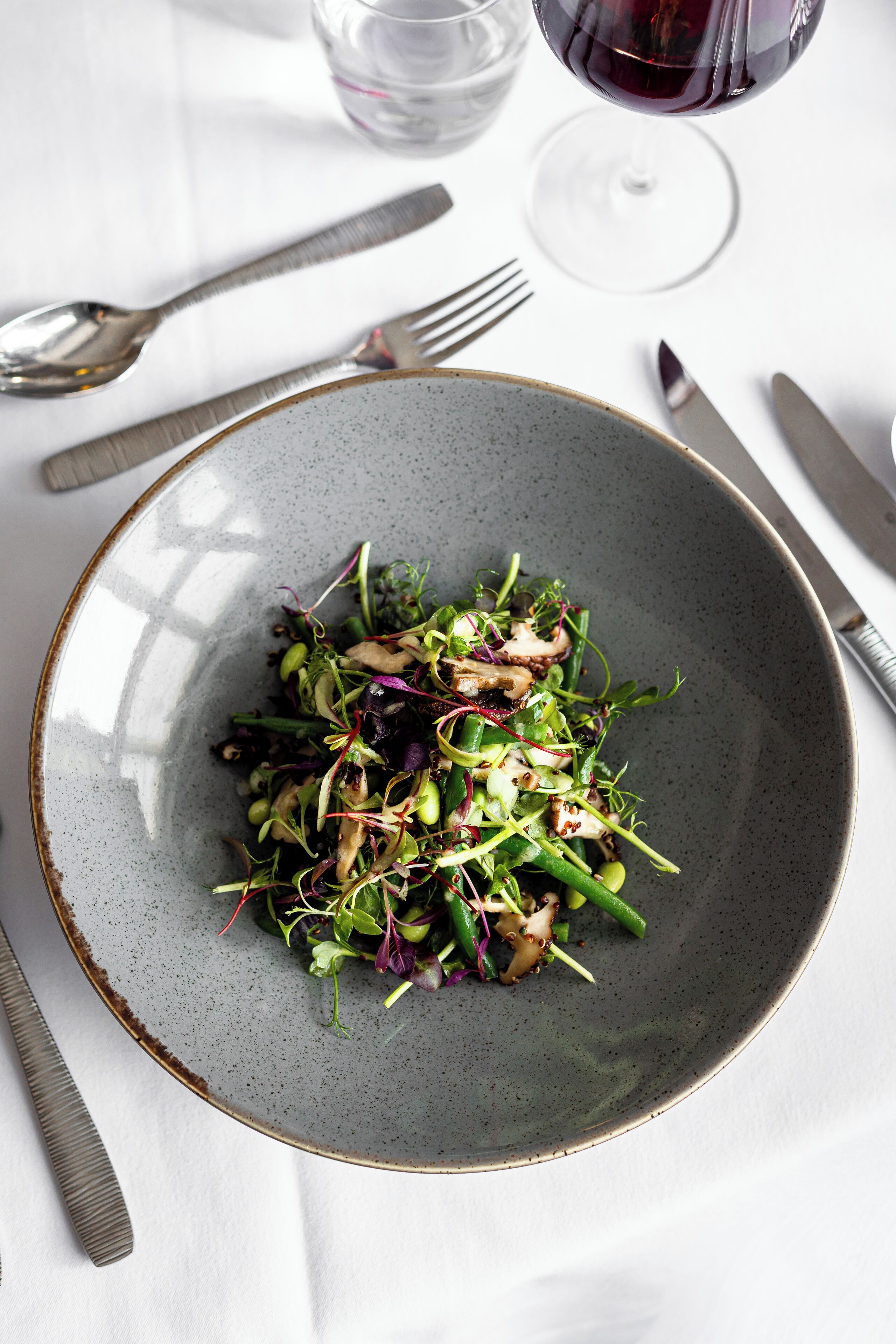 Un plato de ensalada con diferentes verduras y hongos, acompañado de cubiertos y una copa de vino tinto sobre una mesa blanca.