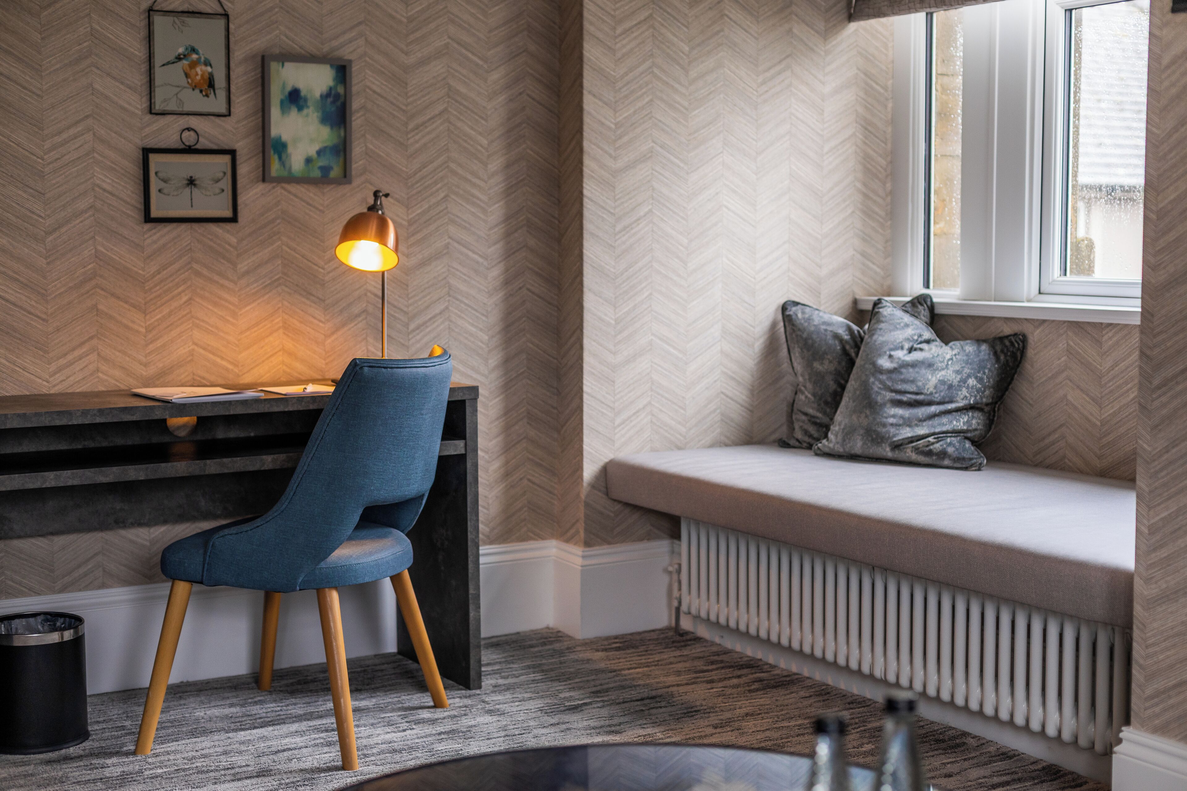 A modern room with a herringbone-patterned wall, a desk with a lamp, a blue chair, a cushioned bench with pillows, and a radiator.