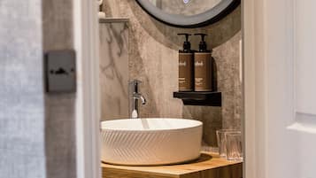 Un baño con un espejo redondo, un lavamanos blanco y una estantería de madera con artículos de aseo.