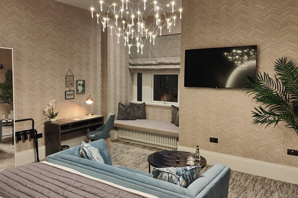 A modern bedroom with a large bed, a desk, a chair, a sofa, a TV, and a large plant.