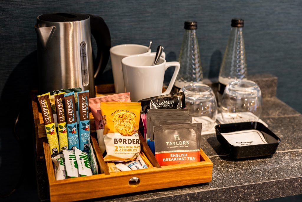 A breakfast tray with tea bags, a coffee pot, mugs, and glass bottles on a countertop.