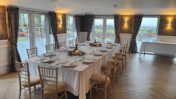 A dining room with a long table set for a meal, large windows with curtains, and a view of the outdoors.