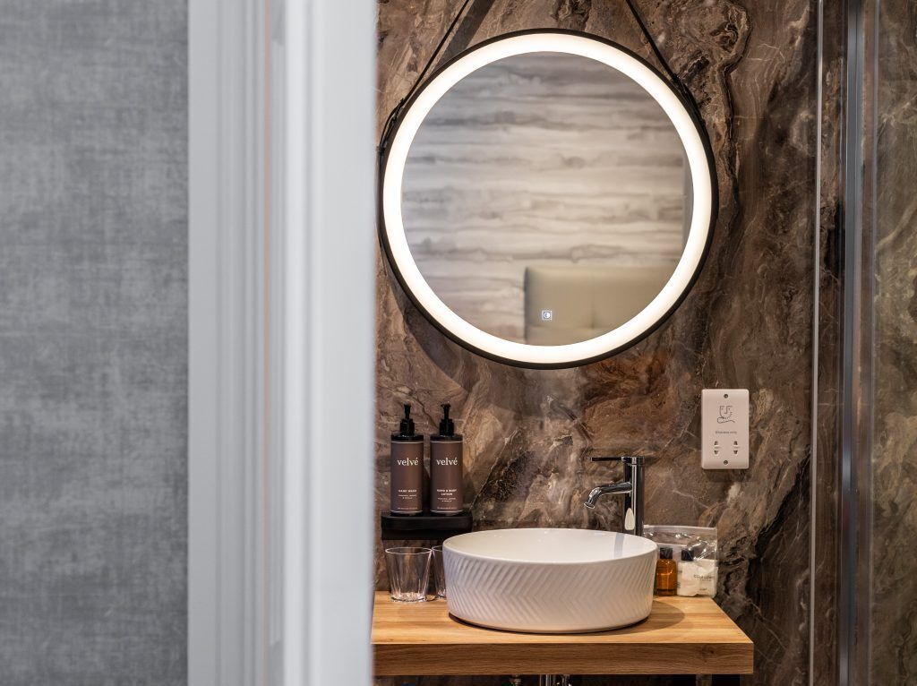 A bathroom with a round mirror, a white sink, and toiletries on a wooden shelf.