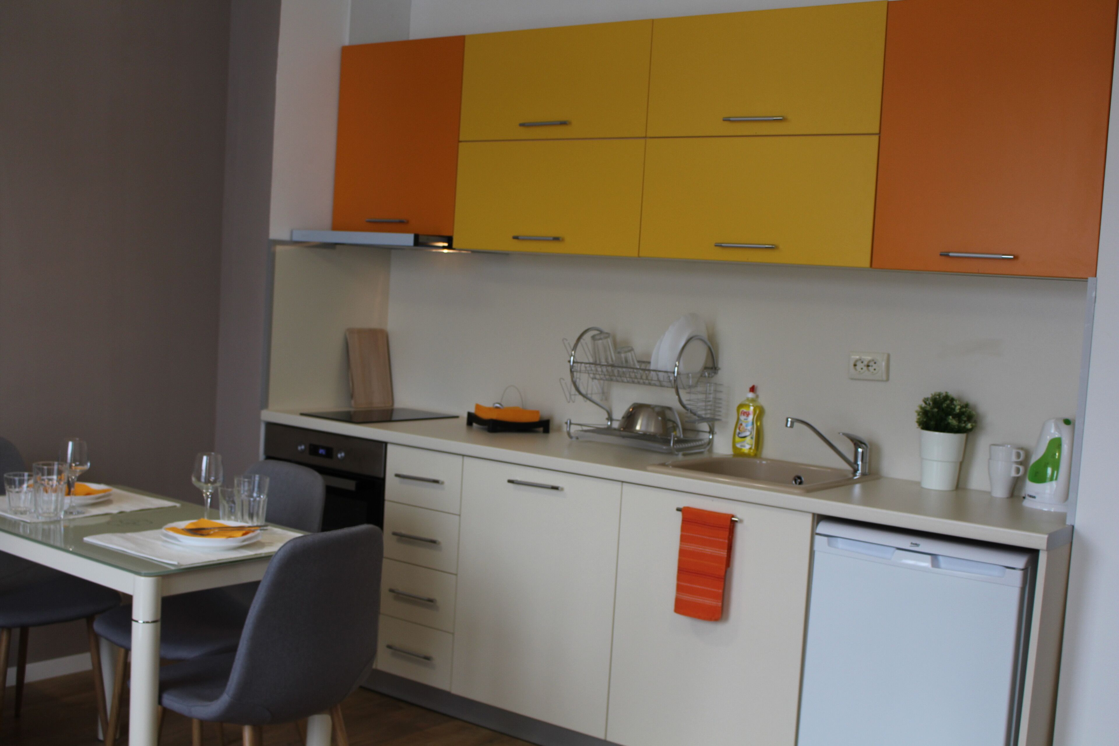 A modern kitchen with white and orange cabinets, a dishwasher, and a dining area with a table set for two.