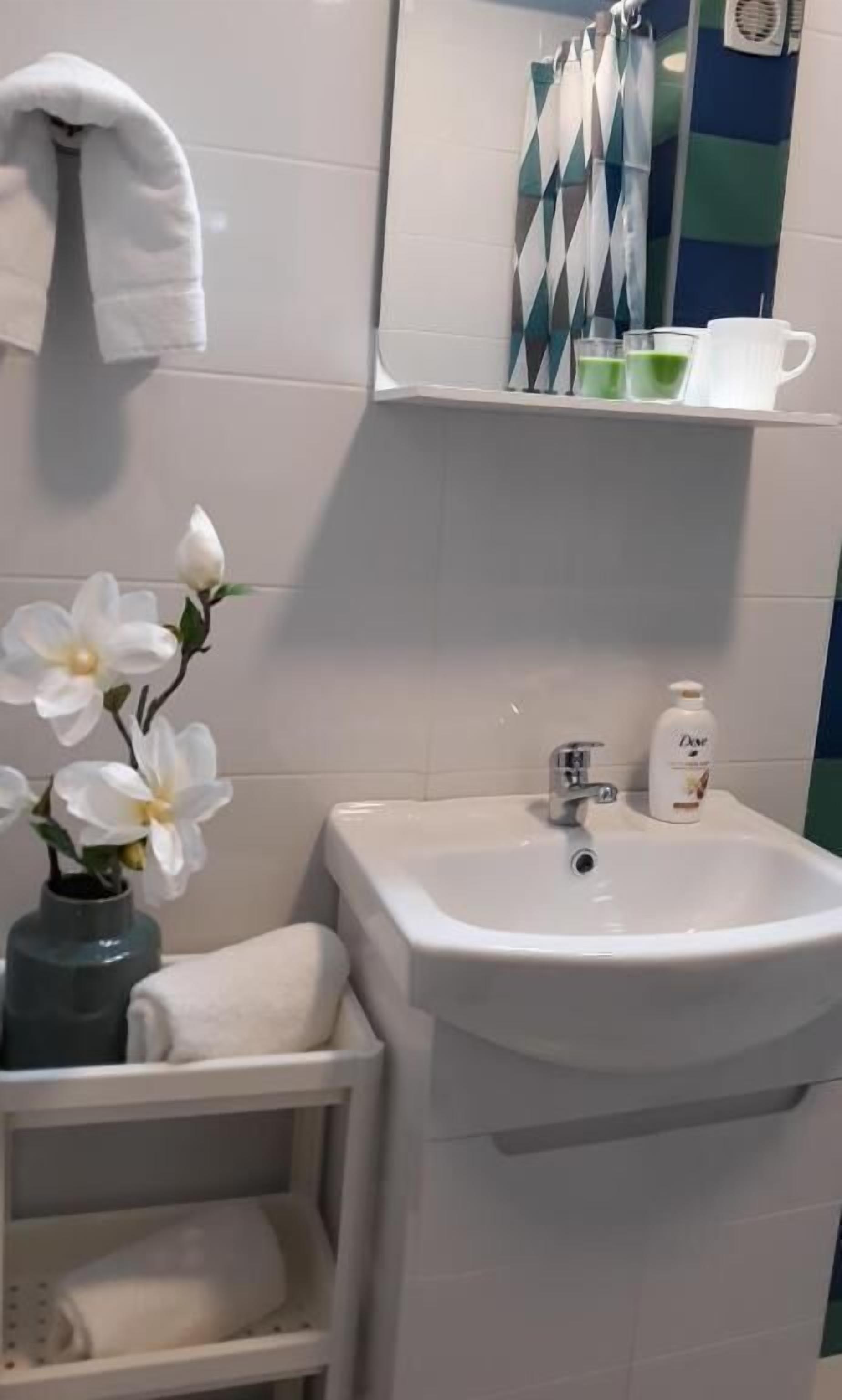 A bathroom with a white sink, a mirror, a towel rack, a shelf with green and white bottles, and a vase with white flowers.