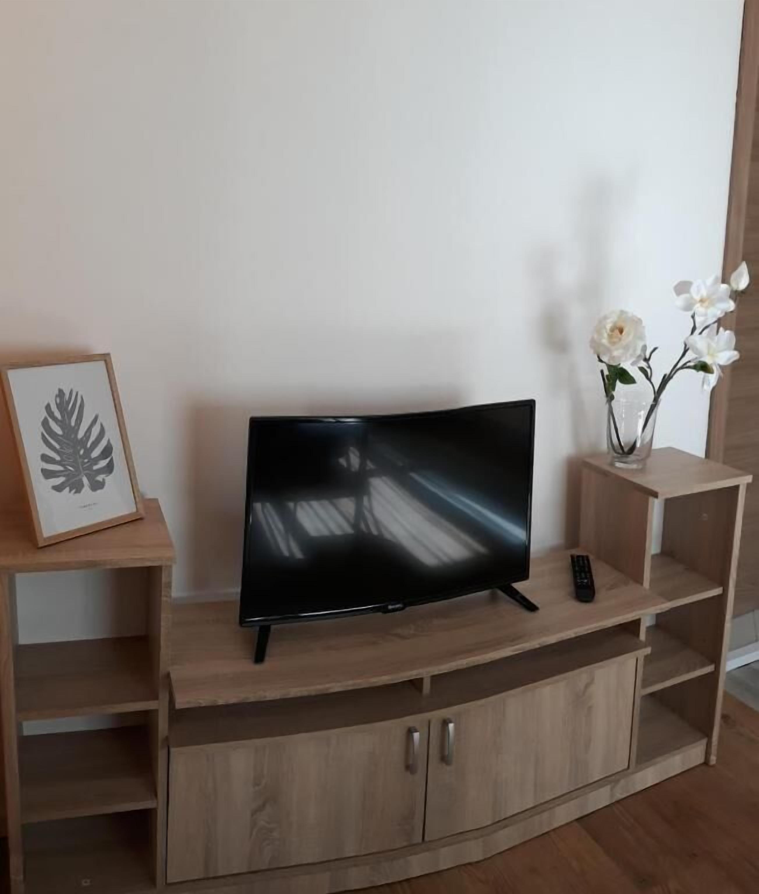 Een flatscreen-tv op een houten dressoir, met een afstandsbediening, een kunstwerk van een blad in een lijst en een vaas met witte bloemen.