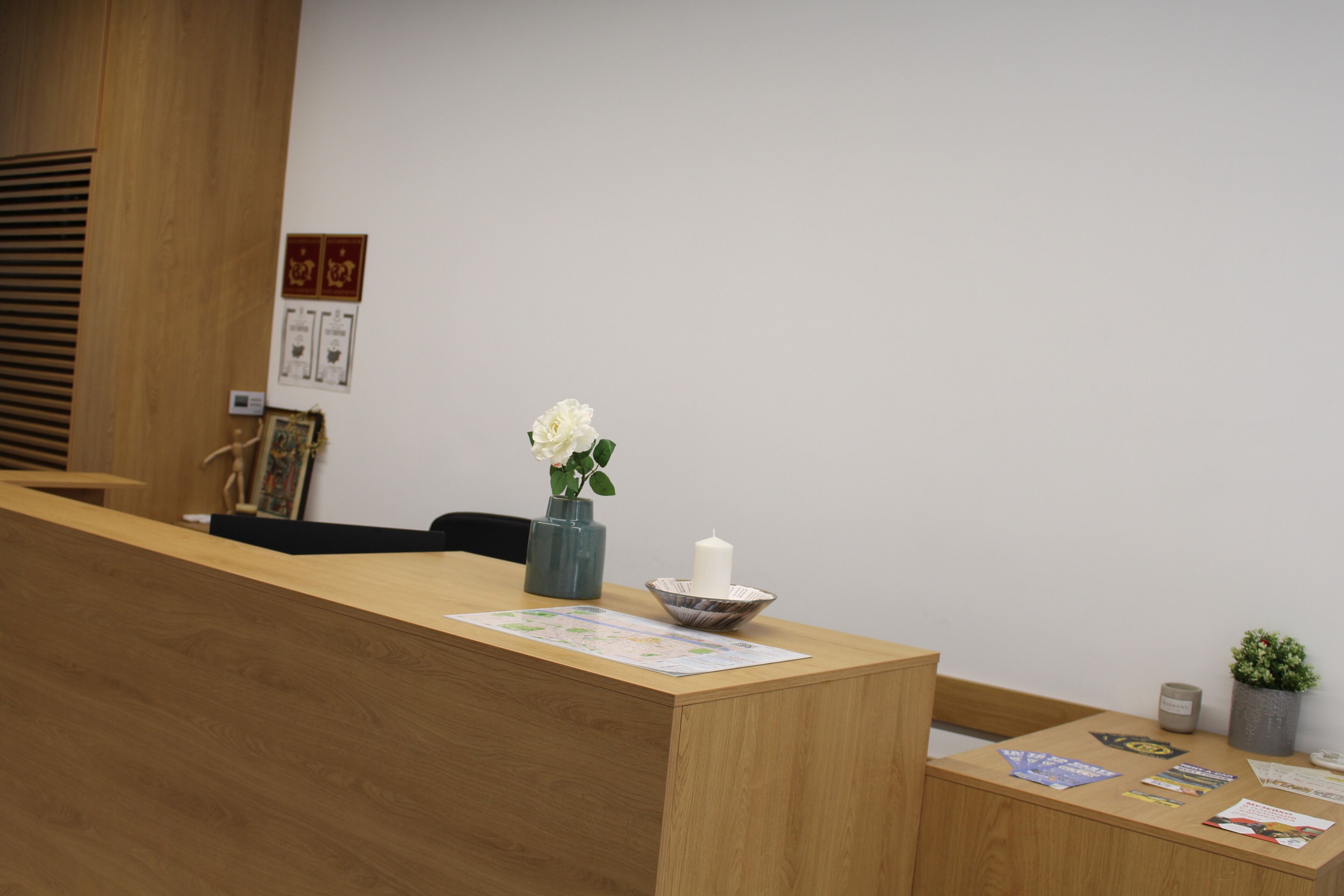 Un mostrador de recepción de madera con un jarrón de flores blancas, una vela y una pequeña planta en maceta. En la pared detrás del mostrador hay diplomas enmarcados.