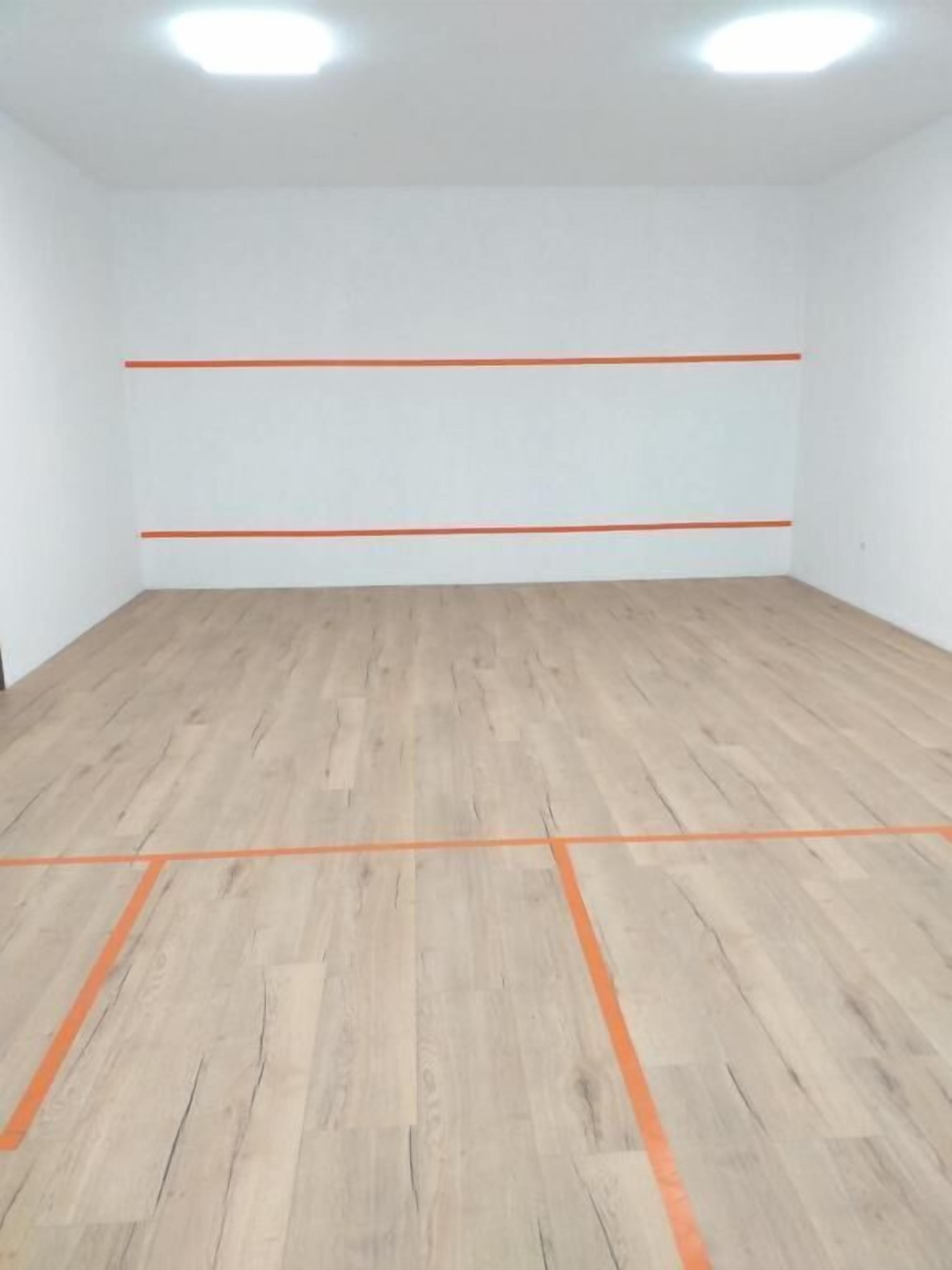 A room with wooden flooring and white walls, marked with orange lines indicating a squash court layout.